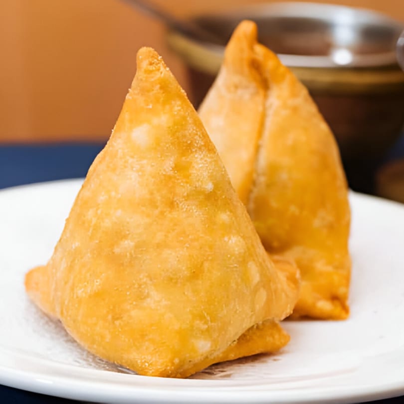 Two golden-brown Indian samosas, triangular and crispy, rest on a white plate.