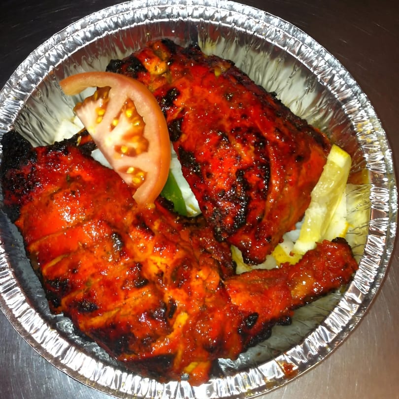 Two pieces of tandoori chicken with a tomato slice and lemon wedges in a foil container.