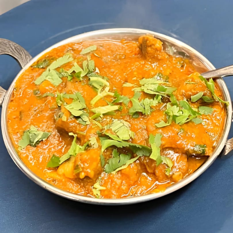 Indian curry with chicken pieces in a rich orange sauce, garnished with fresh cilantro in a metal bowl.