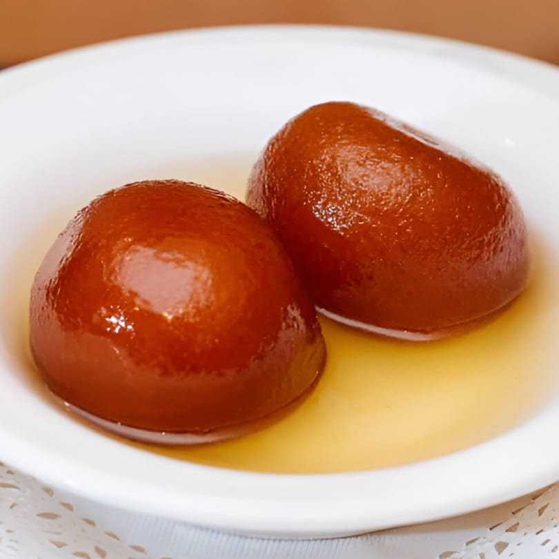Two glistening gulab jamun, Indian fried dough balls, soaked in syrup on a white plate.