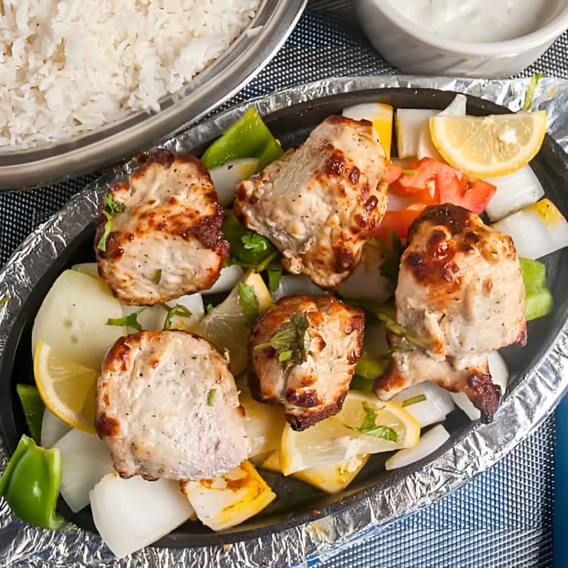 Indian chicken tikka with onions, peppers, and lemon wedges, served with rice and sauce.